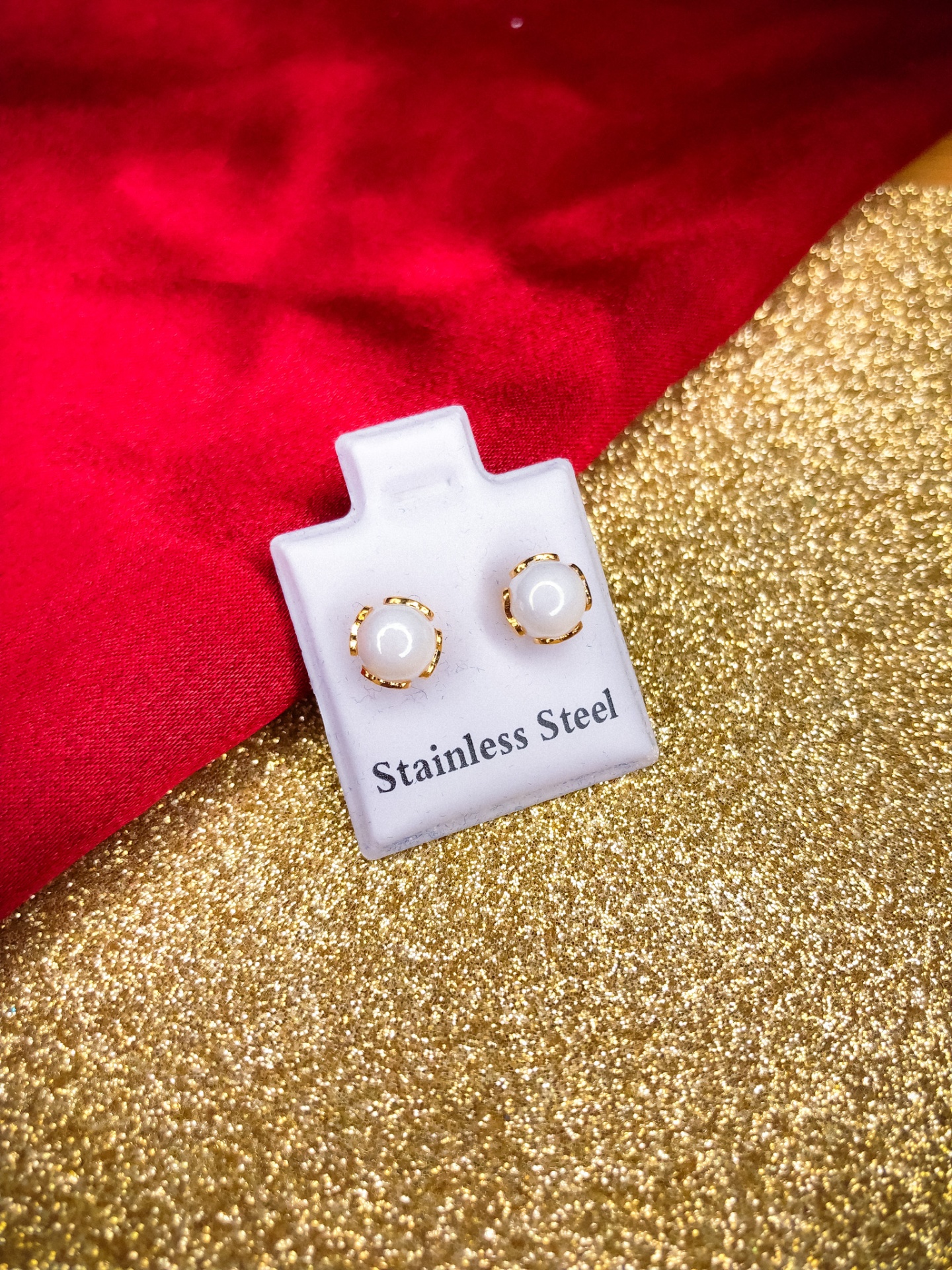 Brincos redondos com pérolas e detalhes dourados em suporte branco sobre fundo dourado e tecido vermelho