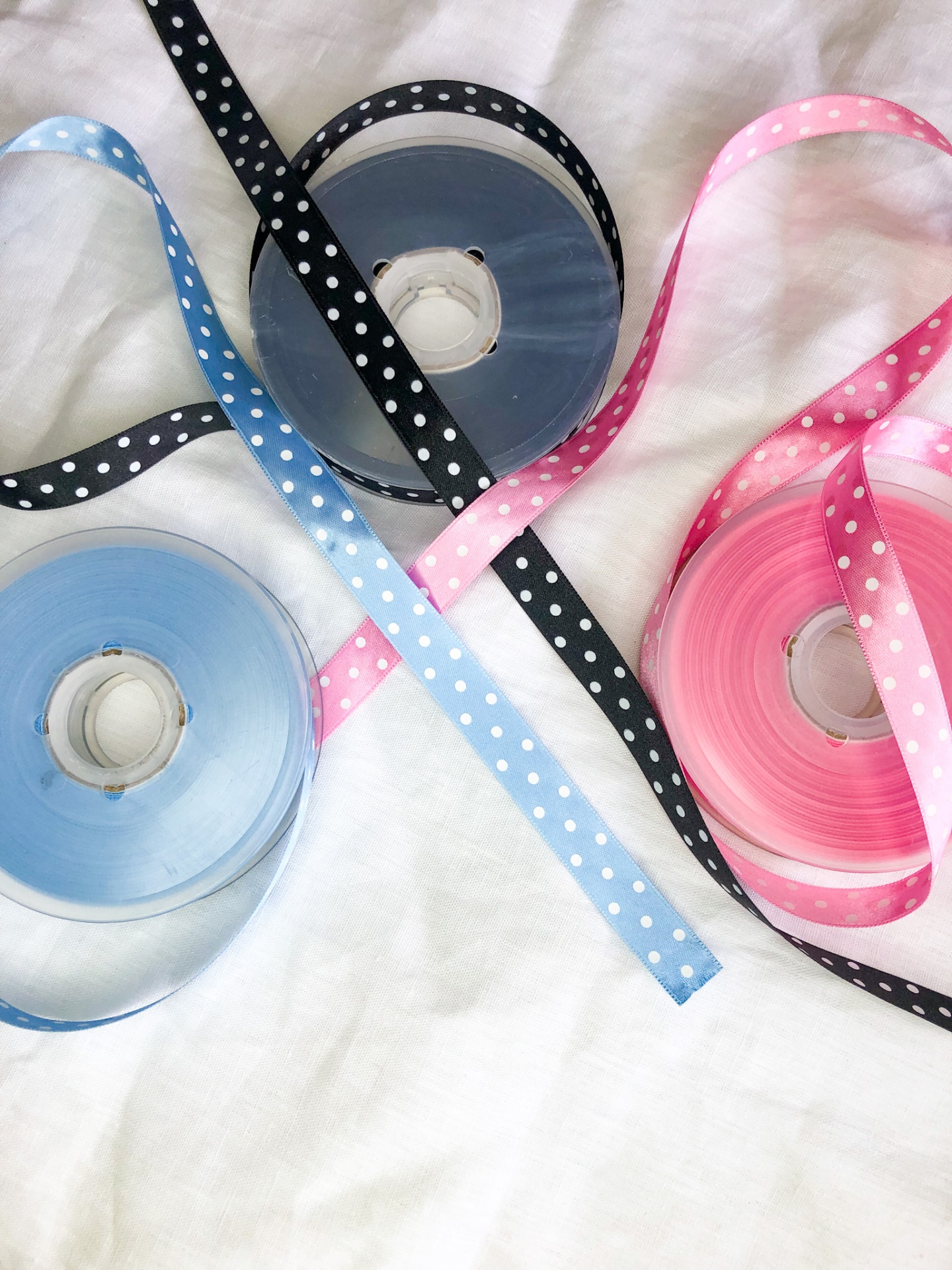 Rolos de fita de cetim azul, preta e rosa com bolinhas brancas sobre tecido branco