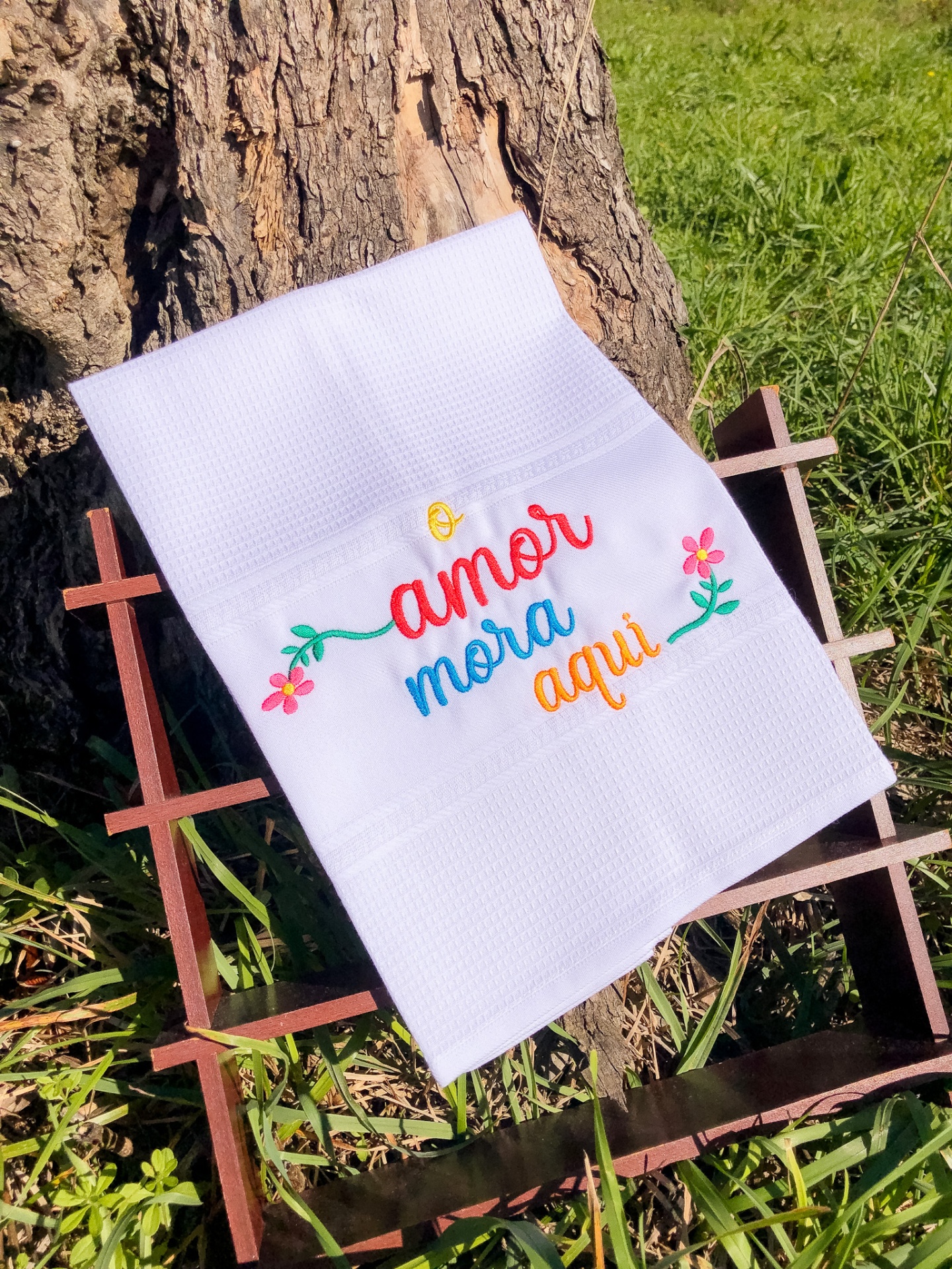 Toalha branca bordada com a frase 'amor mora aqui' e flores, sobre estrutura de madeira junto a um tronco de árvore