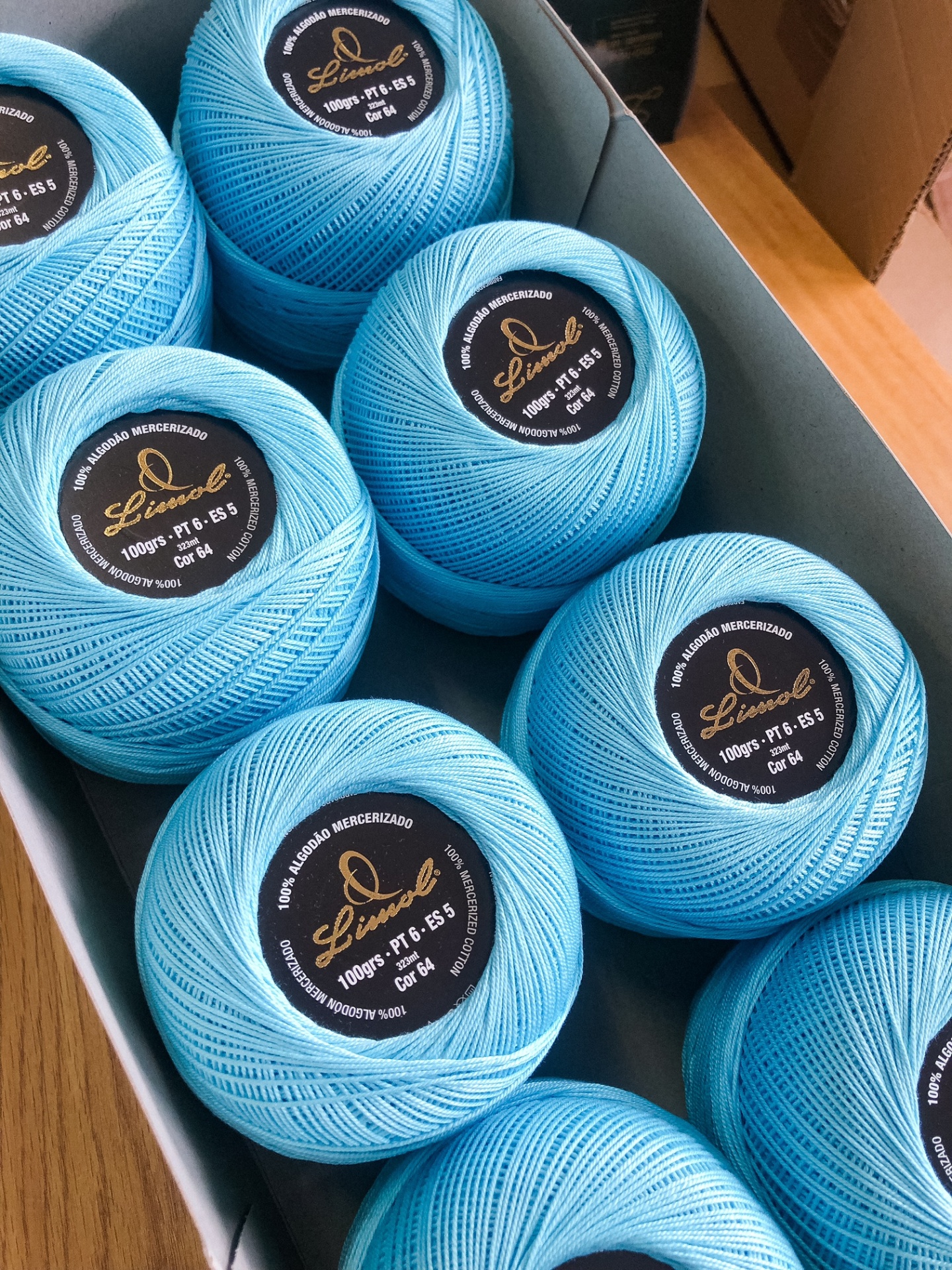 Oito novelos de fio de algodão azul claro em bobinas redondas com etiquetas pretas numa caixa cinza