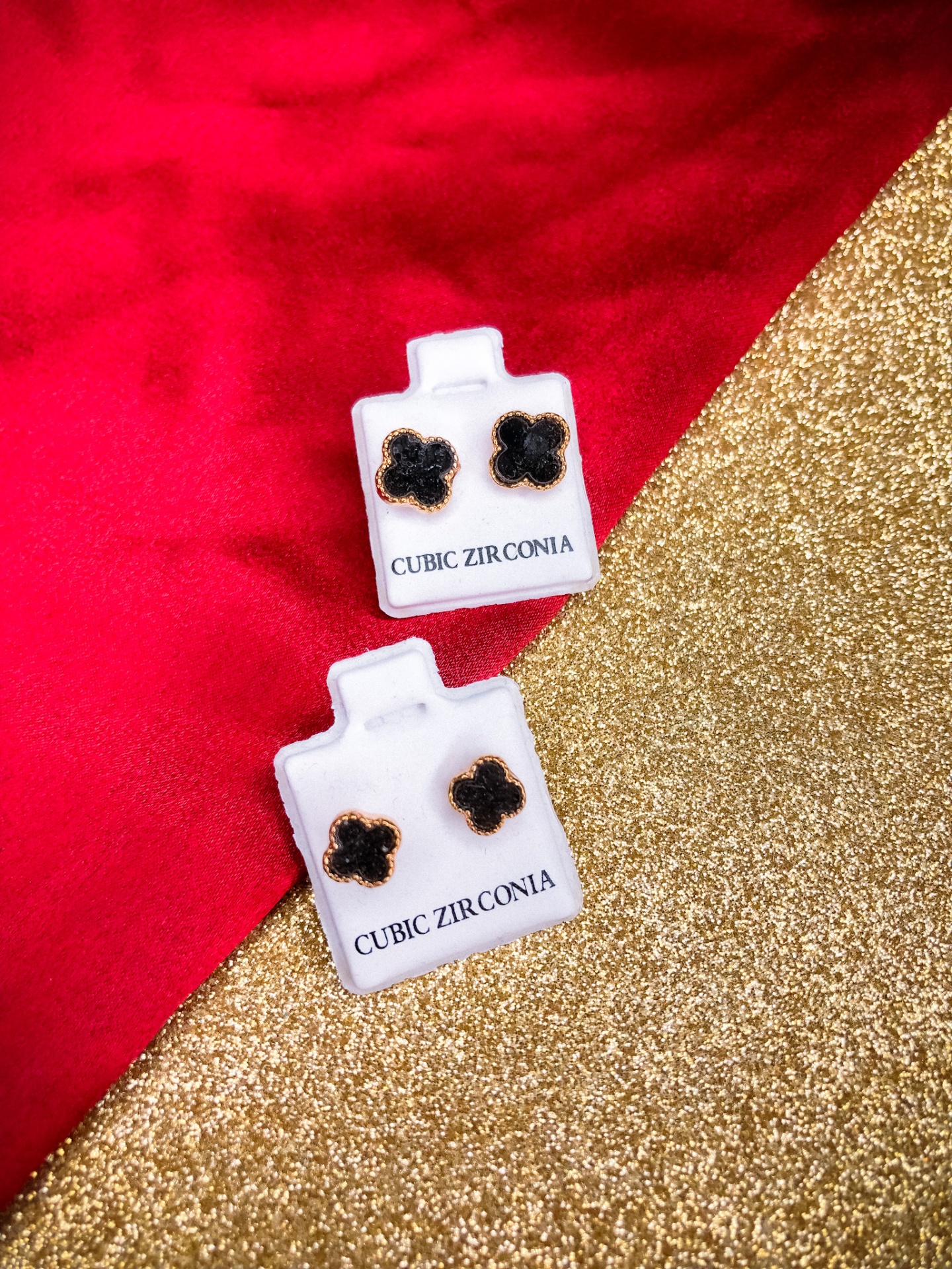 Dois pares de brincos florais pretos e dourados em suportes brancos com texto CUBIC ZIRCONIA, sobre fundo vermelho e dourado