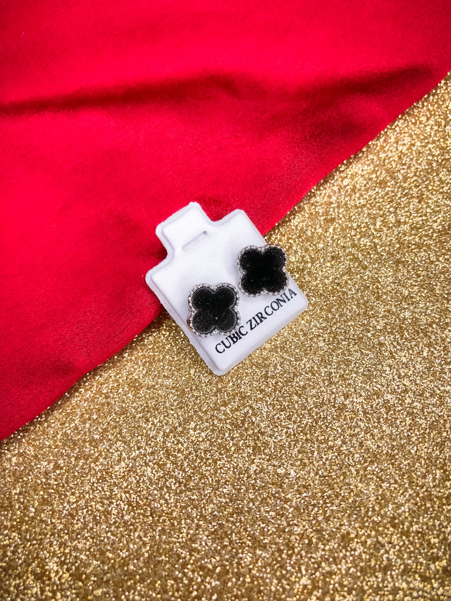 Brincos florais pretos em suporte branco com texto 'CUBIC ZIRCONIA' sobre fundo dourado e vermelho