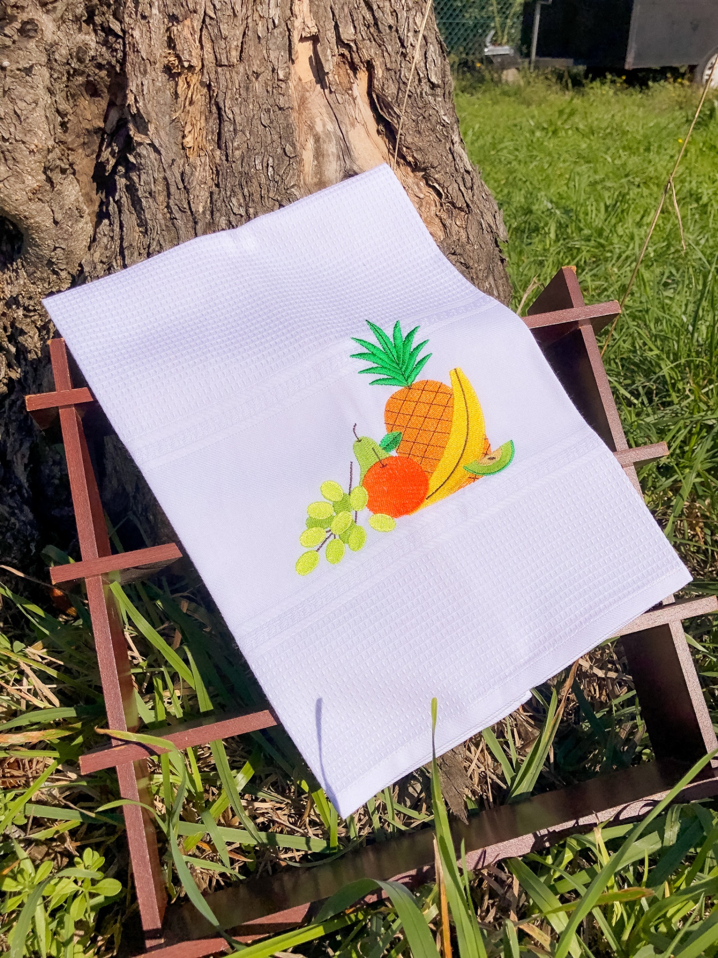 Toalha branca com bordado de frutas pendurada numa estrutura de madeira ao ar livre