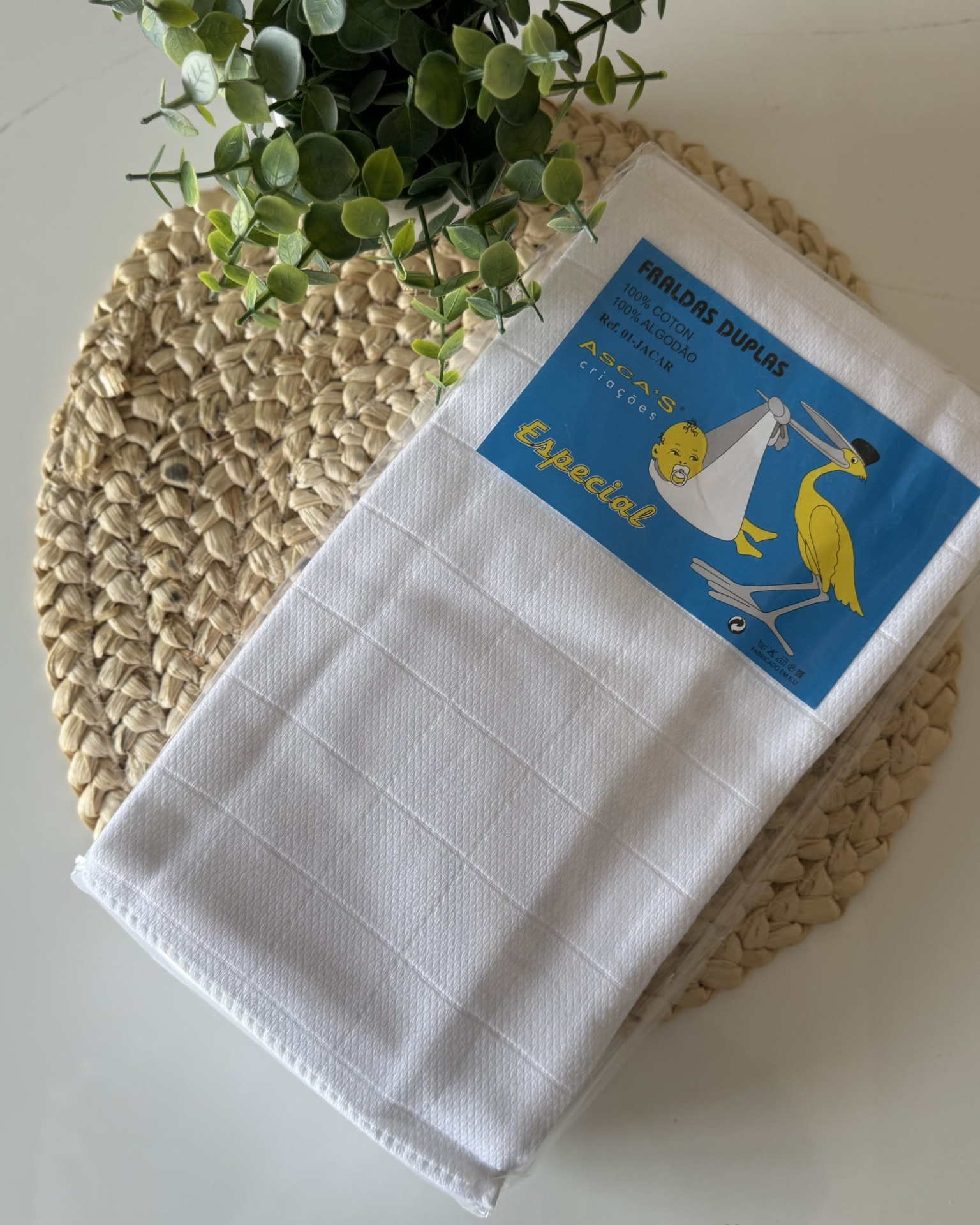 Fralda dupla branca com etiqueta azul e ilustração sobre esteira de fibras naturais