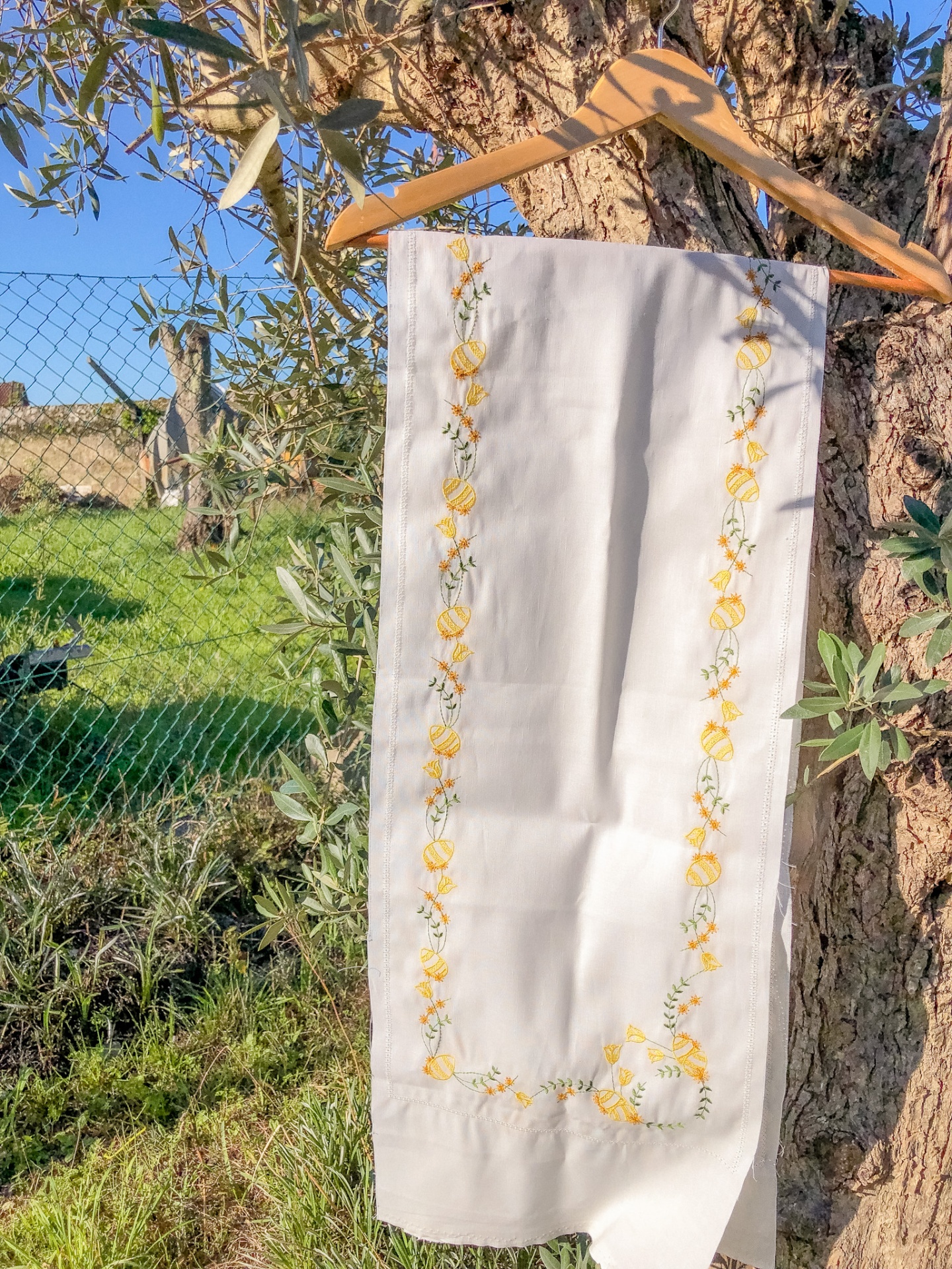 Toalha branca com bordados amarelos e verdes pendurada num cabide numa árvore