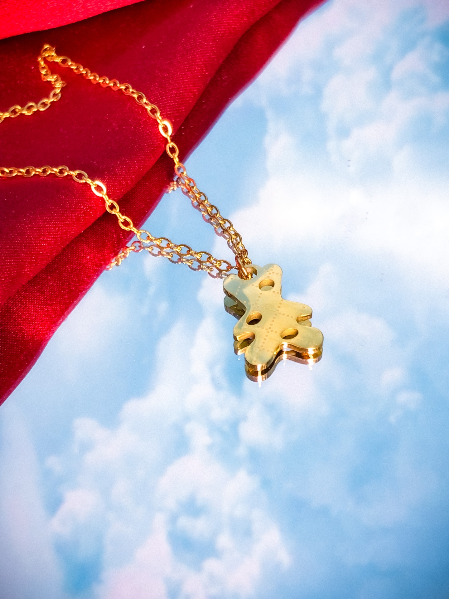 Colar dourado com pendente em forma de folha sobre fundo vermelho e espelho refletindo céu