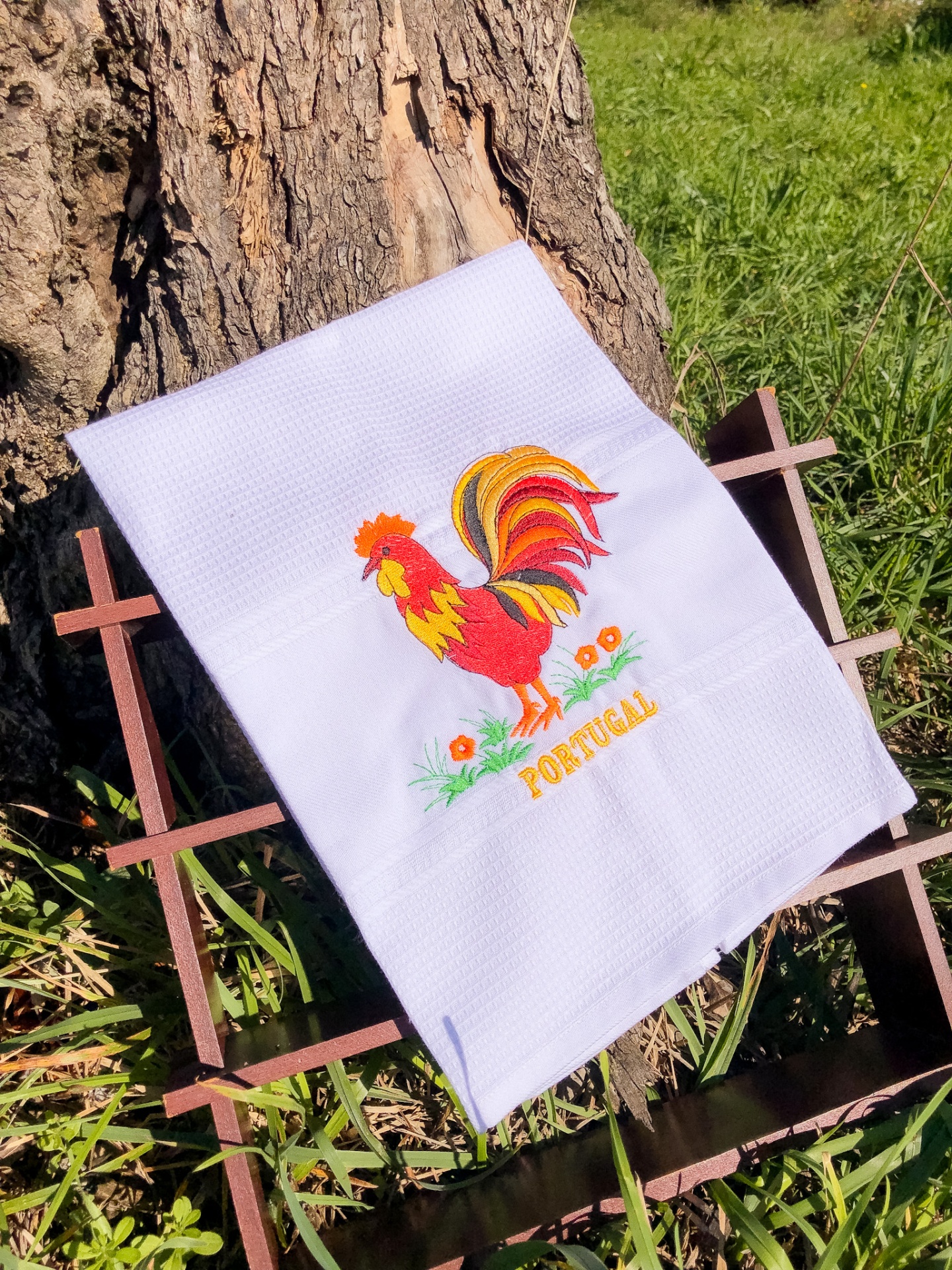 Toalha branca bordada com galo colorido e texto PORTUGAL sobre estrutura de ferro ao lado de árvore