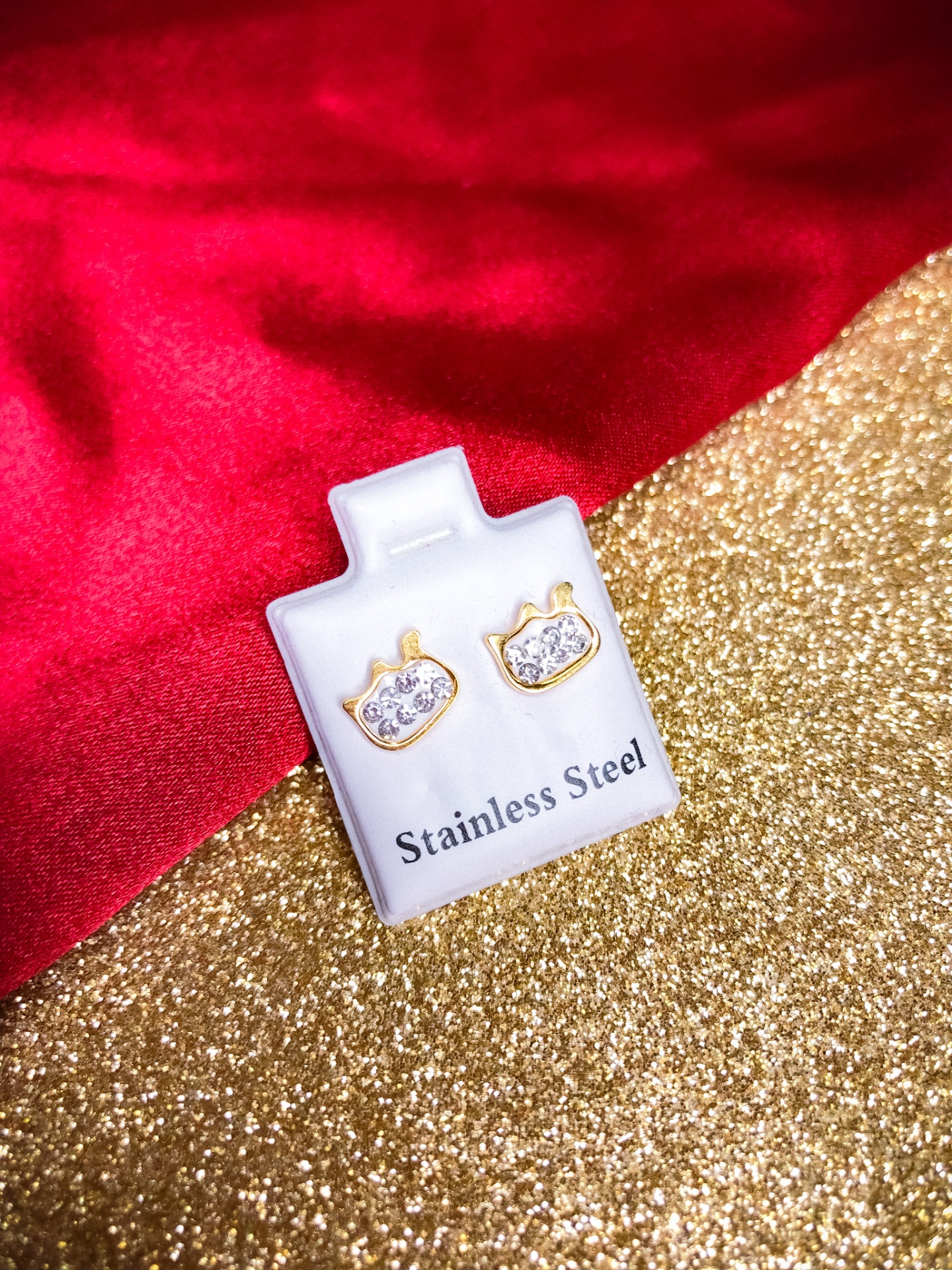 Brincos em aço inoxidável dourados com pedras brilhantes num cartão branco com texto, sobre pano vermelho e fundo dourado
