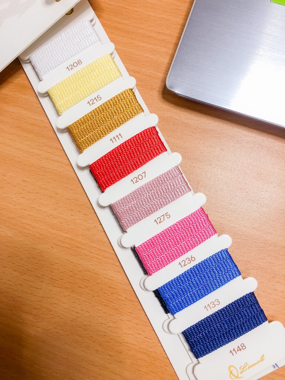 Mostruário de fitas têxteis coloridas em várias cores e etiquetas com números numa mesa de madeira.