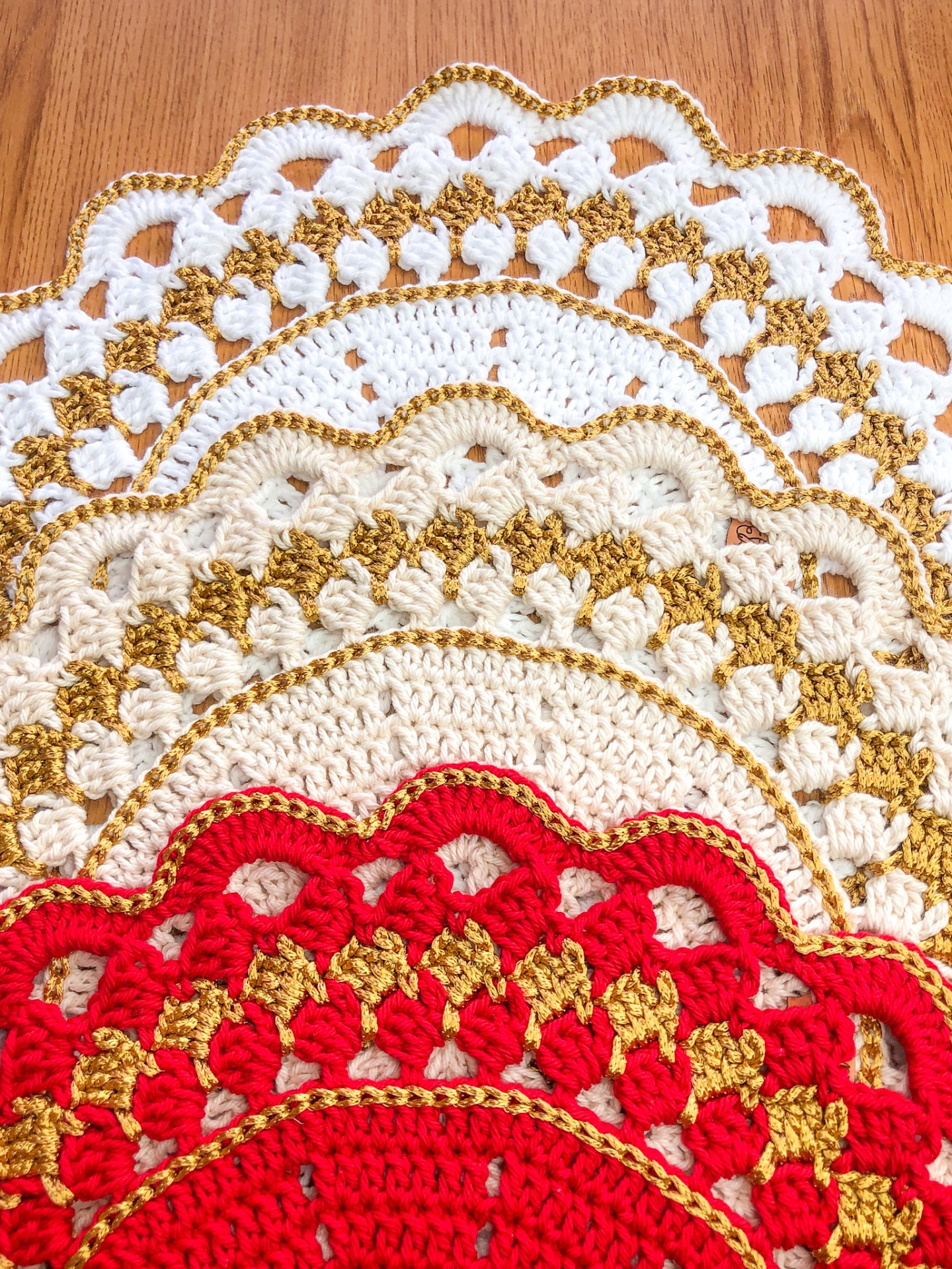 Toalhas de crochê redondas com bordas onduladas coloridas em branco, bege, dourado e vermelho em madeira clara.