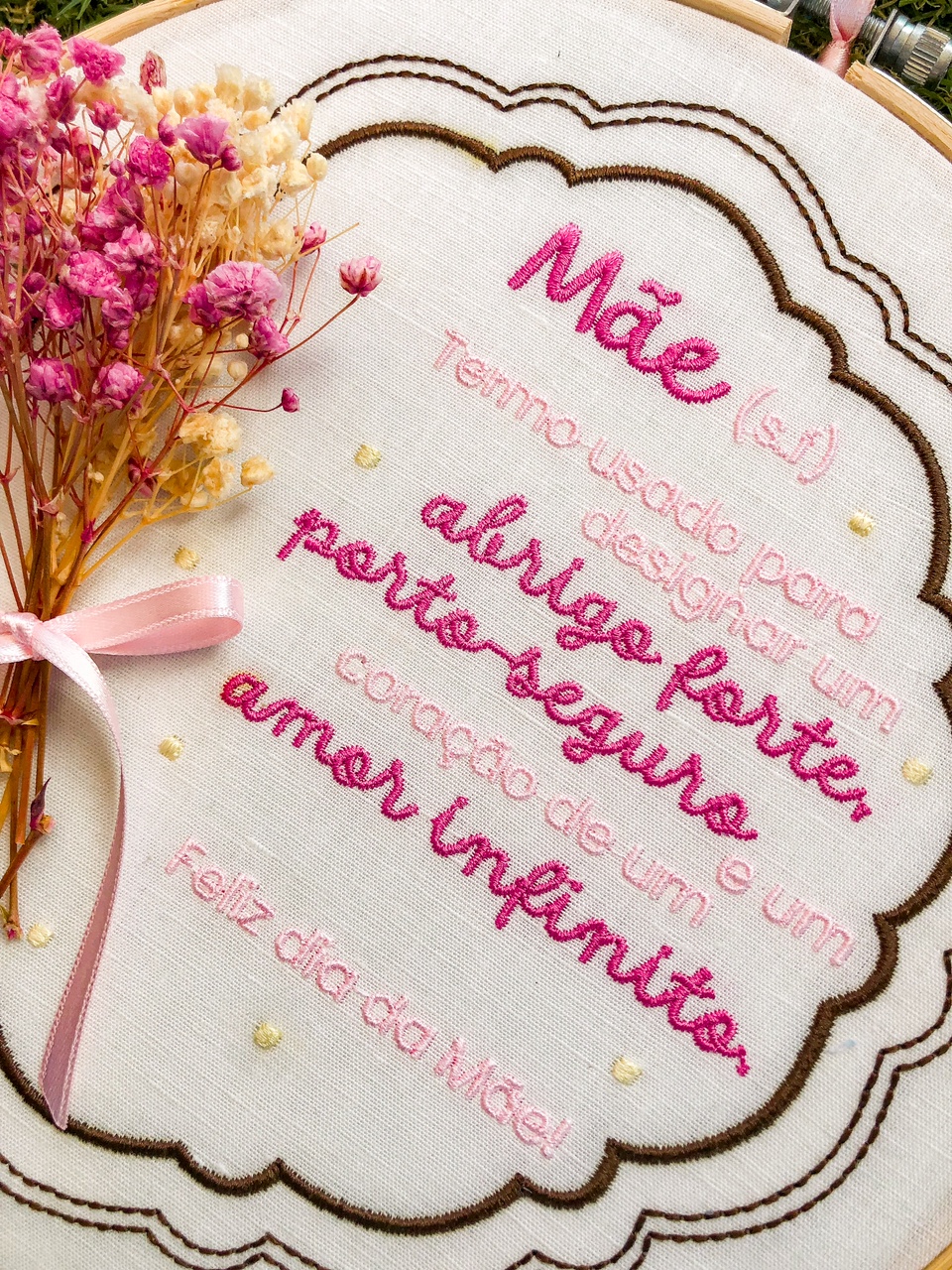 Bordado em tecido branco com texto em rosa e vermelho e ramo de flores secas com fita rosa.