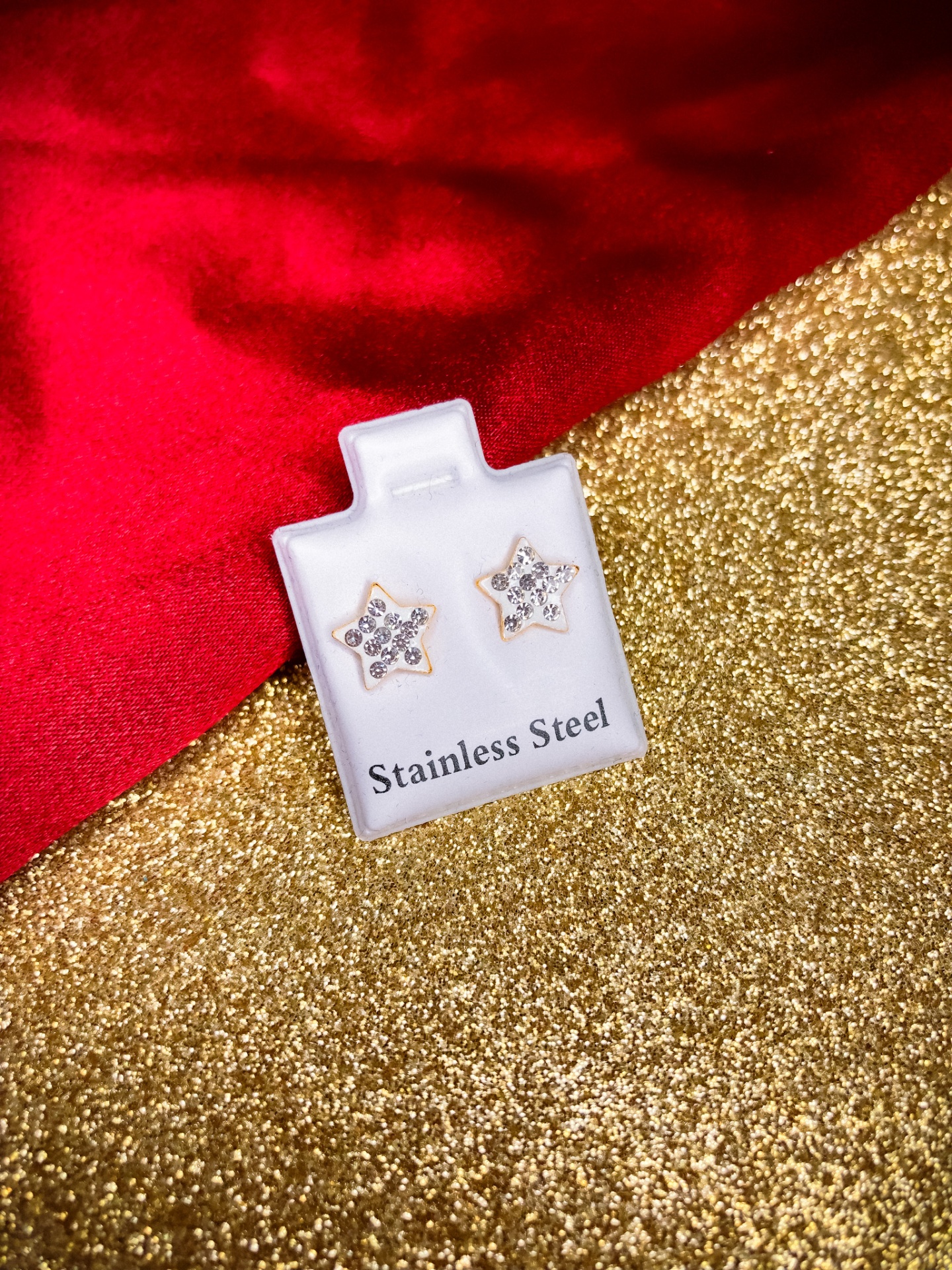 Brincos de estrela em aço inoxidável com pedras brilhantes em embalagem branca sobre fundo dourado e vermelho
