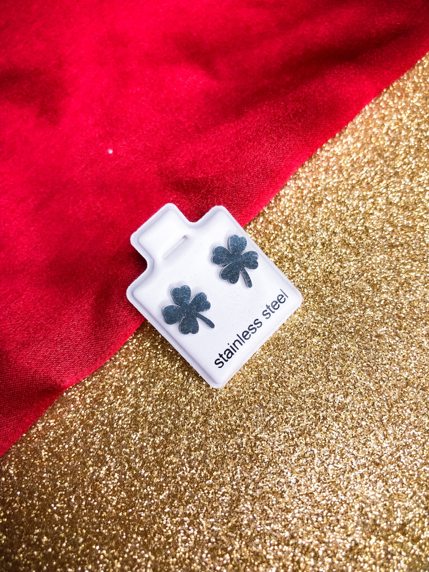 Brincos de aço inoxidável em forma de trevo em suporte branco com texto, sobre fundo vermelho e dourado