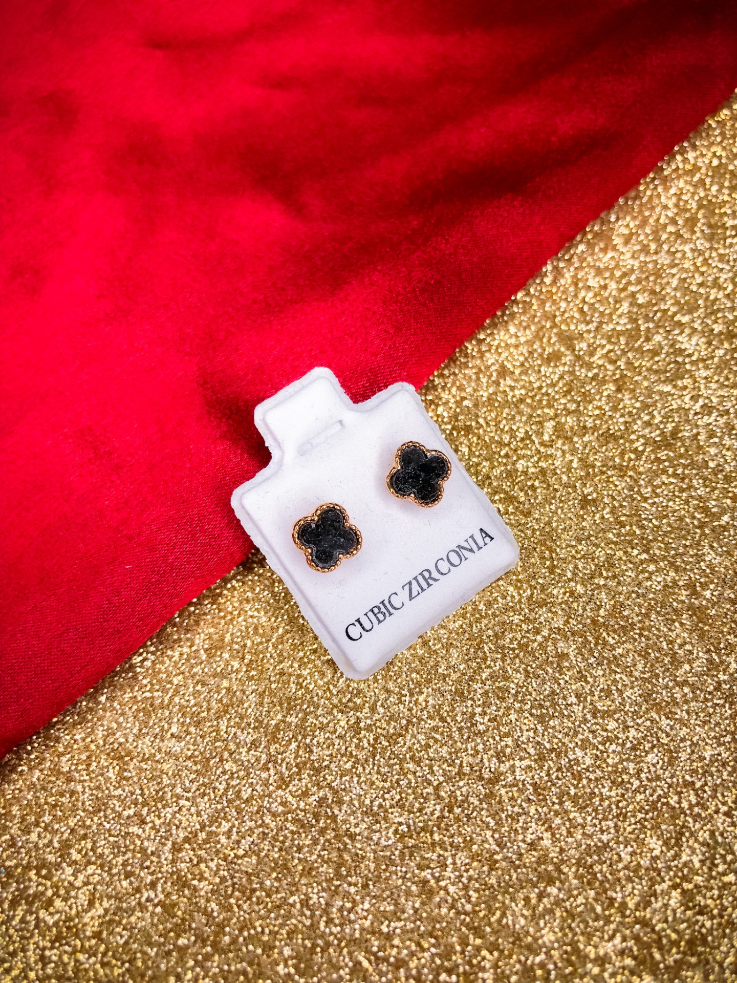 Brincos trevo preto em cartão branco com texto Cubic Zirconia em fundo dourado brilhante e tecido vermelho.