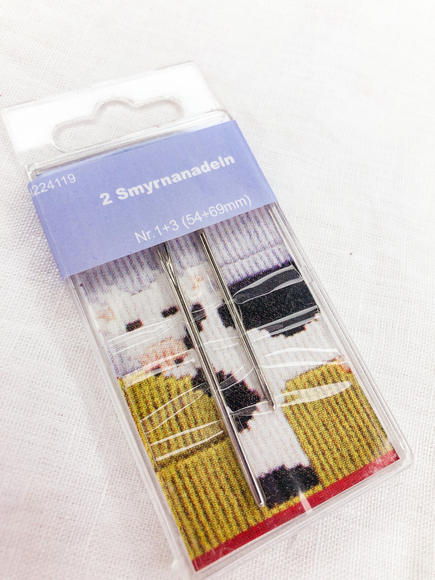 Embalagem de agulhas Smyrnanadeln de metal com cartão azul e padrão listrado colorido.