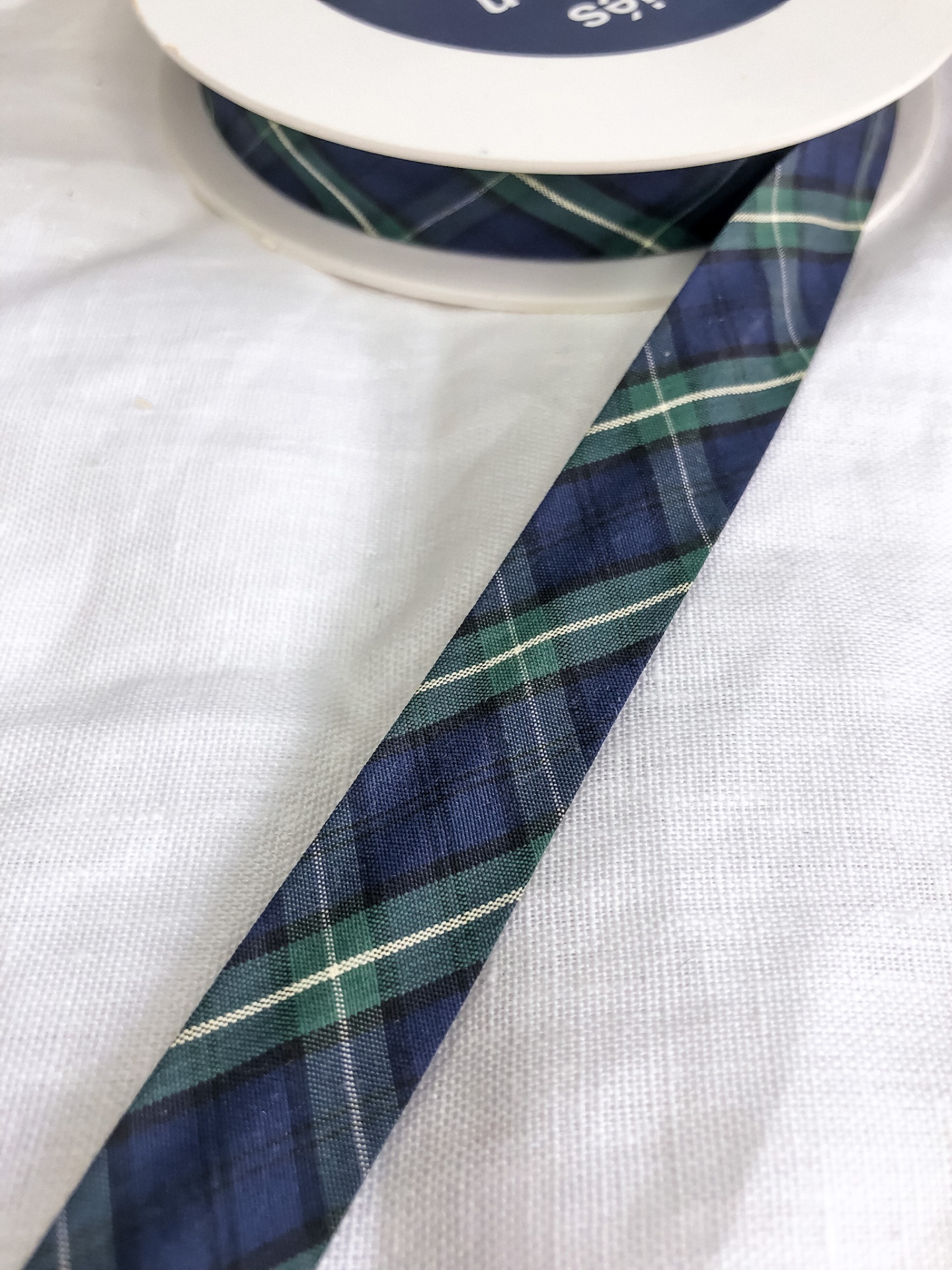 Fita xadrez azul, verde e branco enrolada em carretel branco sobre tecido branco.