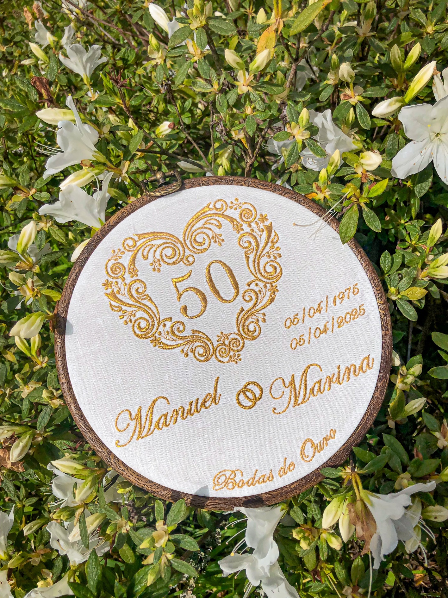 Bordado redondo branco com design dourado de coração e texto comemorativo rodeado de folhagem e flores brancas