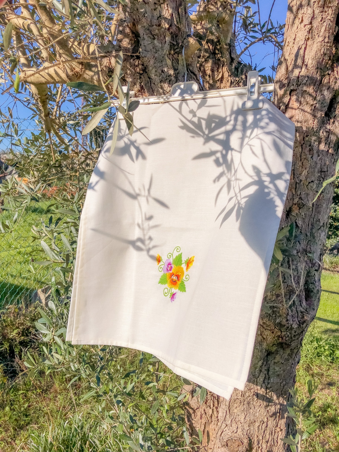 Toalha branca bordada pendurada em árvore com sombra e céu azul