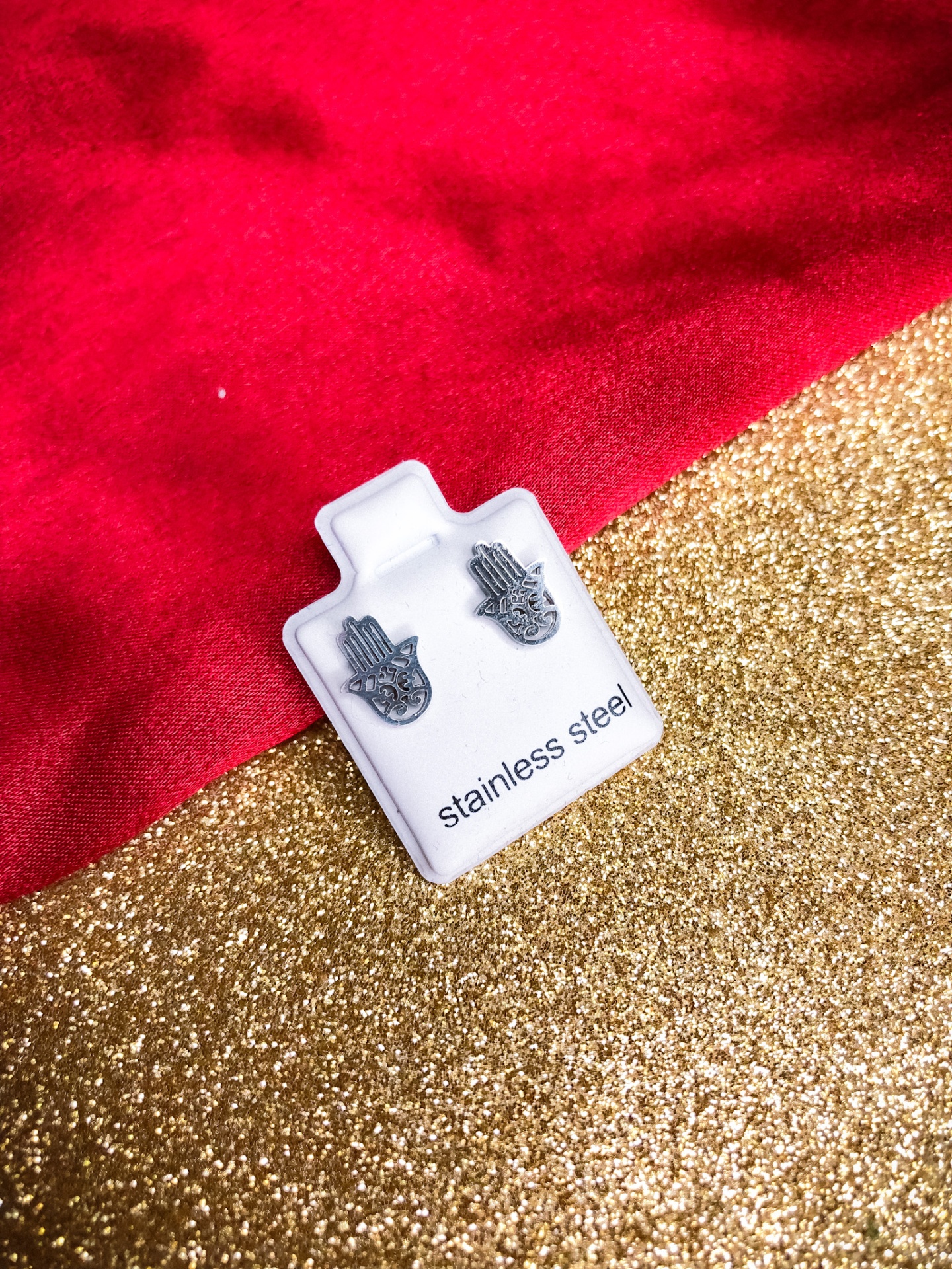 Brincos de aço inoxidável em forma de mão em suporte branco sobre fundo vermelho e dourado