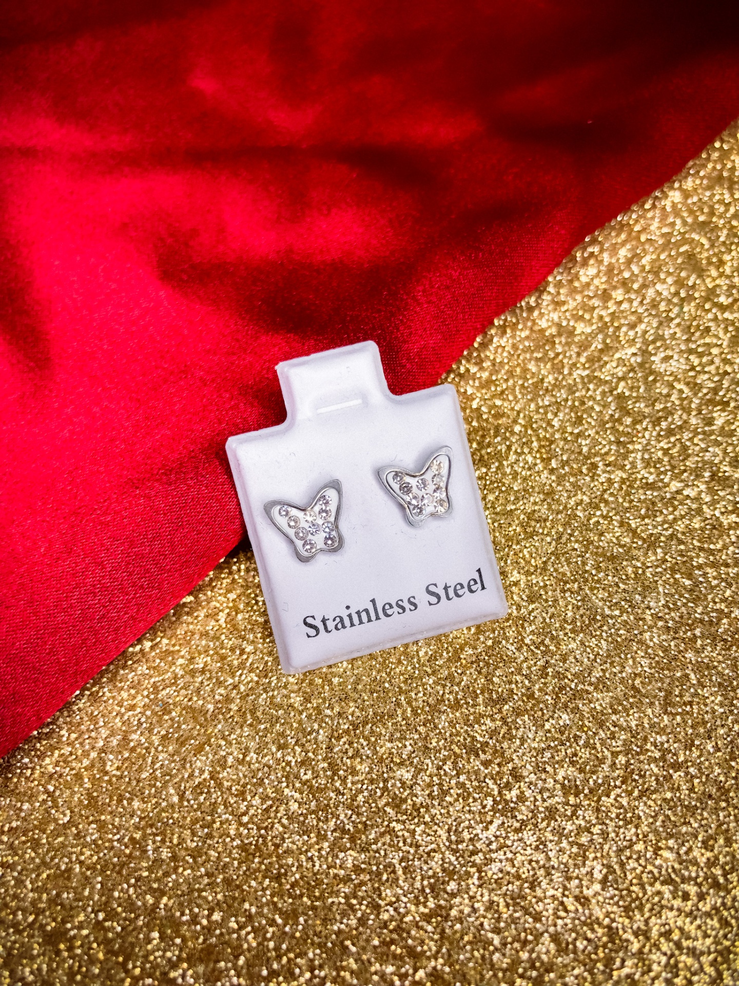 Brincos de borboleta em aço inoxidável com cristais sobre base branca e fundo dourado e vermelho