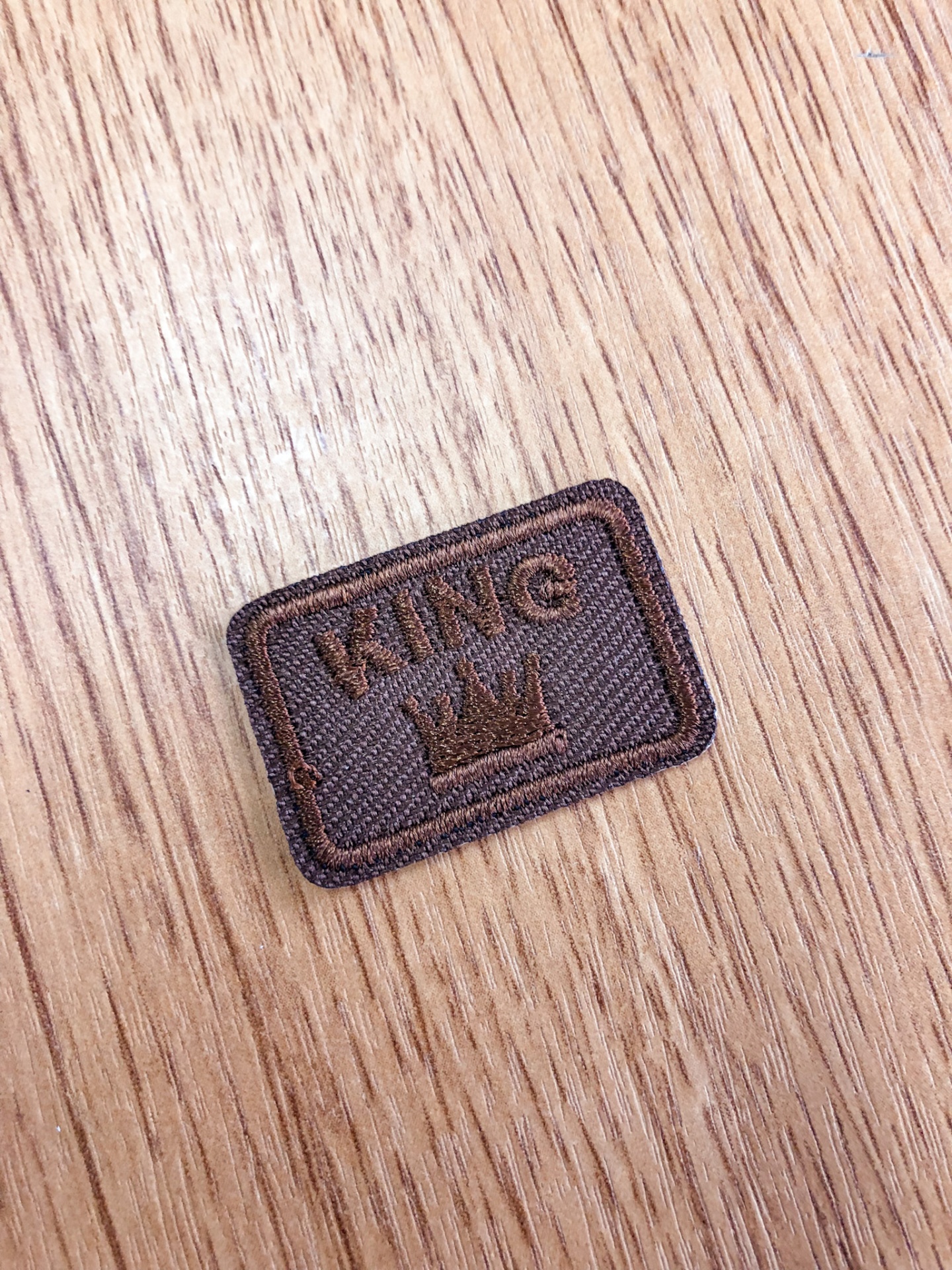 Etiqueta retangular de tecido castanho com texto KING e coroa bordados