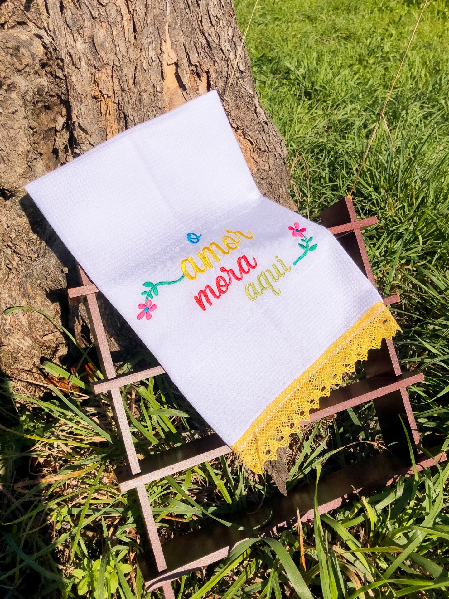 Toalha branca bordada com frase colorida e renda amarela sobre estrutura de madeira na natureza
