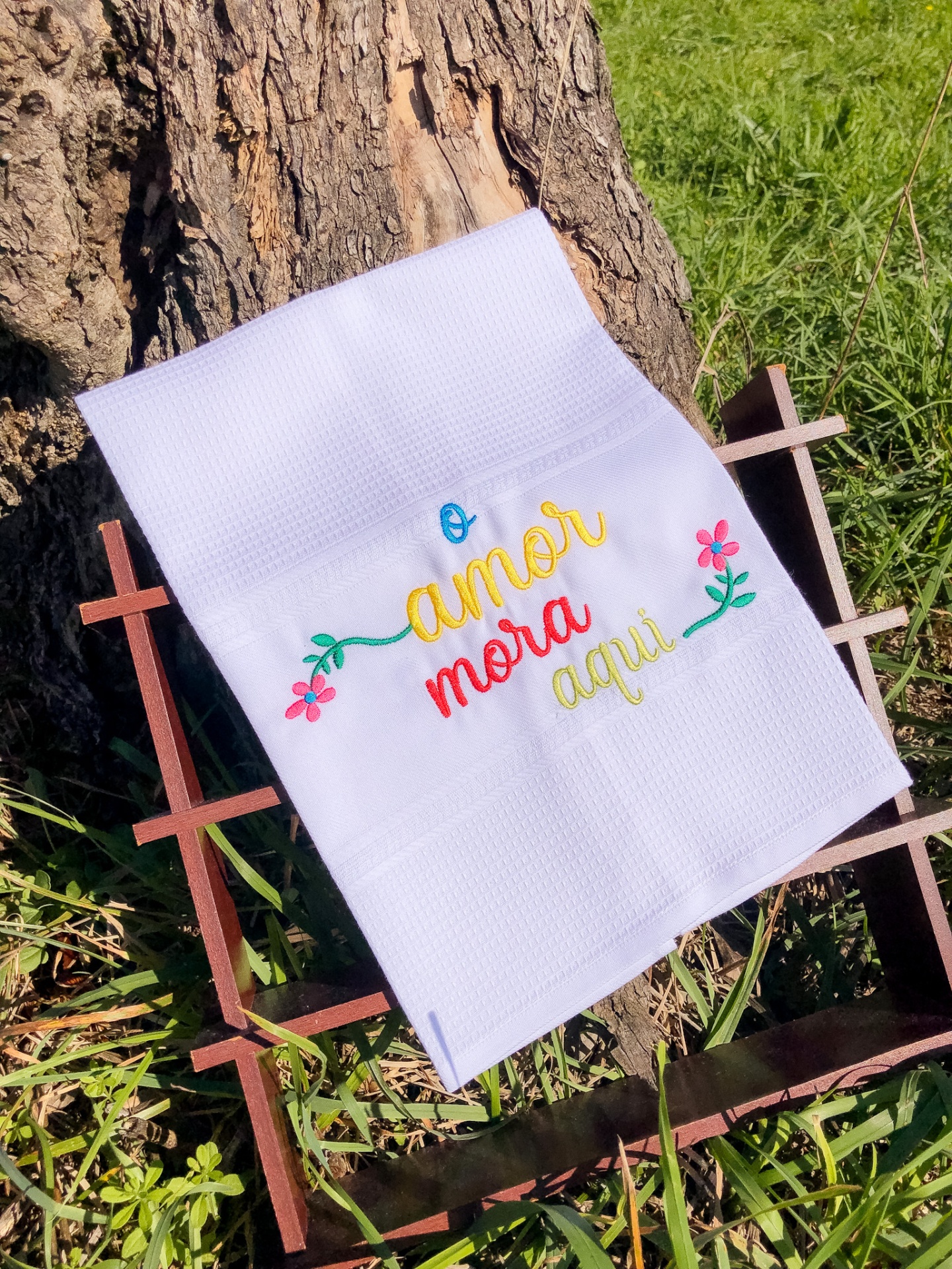 Toalha branca com bordado colorido 'o amor mora aqui' apoiada em estrutura de madeira frente a tronco de árvore