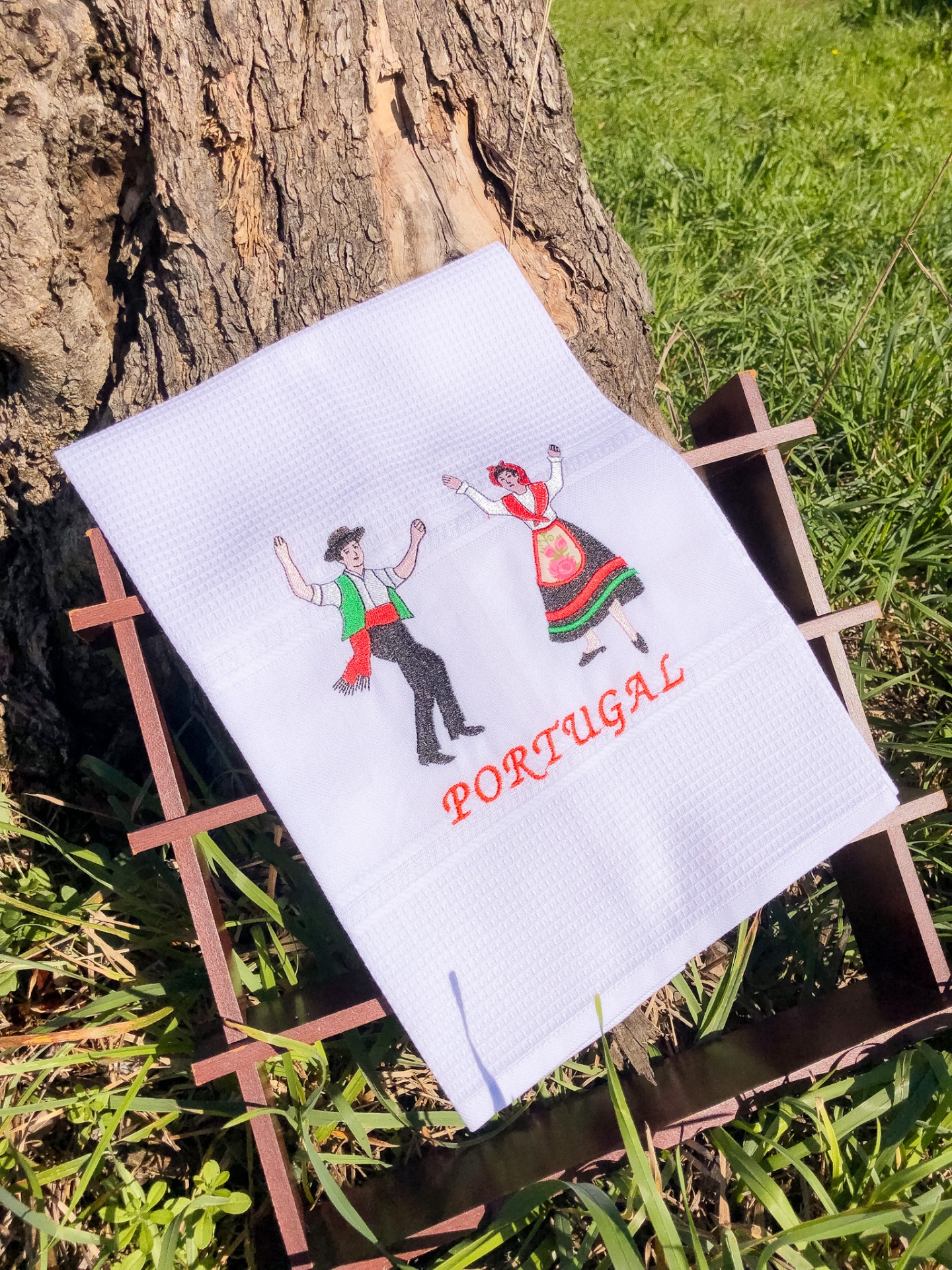 Toalha branca bordada com casal tradicional português e texto PORTUGAL sobre suporte de madeira junto a árvore.