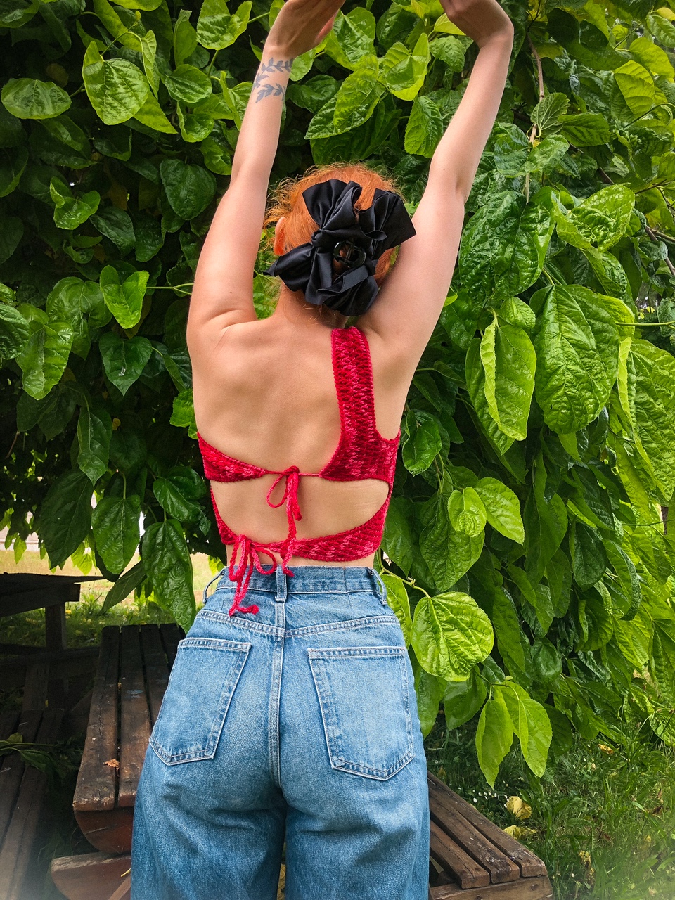 Mulher de costas com cropped vermelho e jeans azul, cabelo preso com scrunchie preto, em frente a folhagem verde.