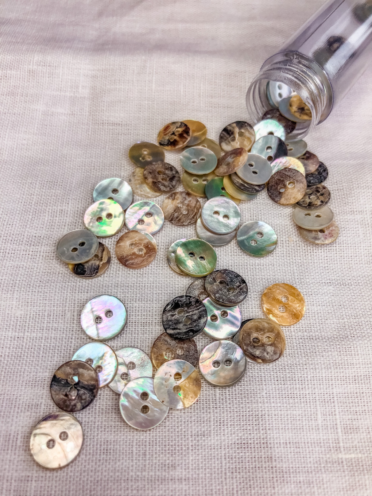 Botões redondos de vários tons e texturas sobre tecido bege claro, com frasco transparente ao lado