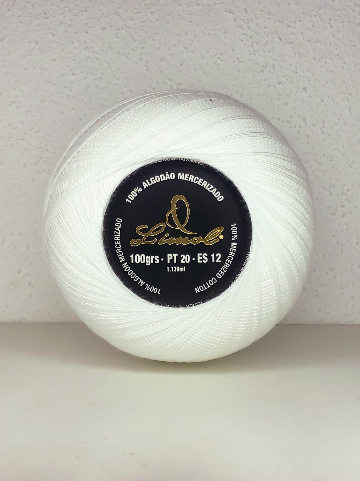 Novelo de linha branca com rótulo preto e letras douradas sobre fundo branco texturado