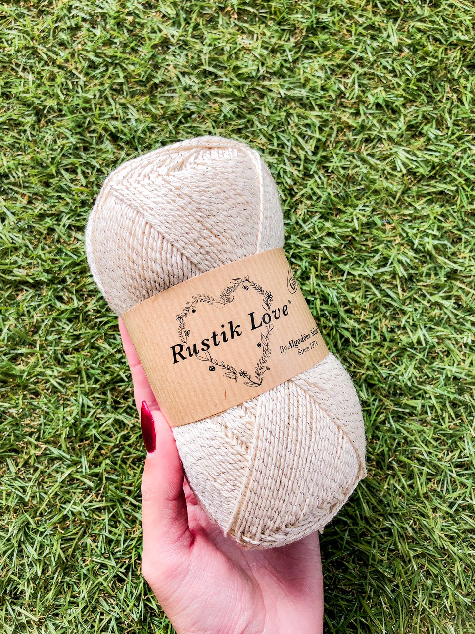 Skein de fio bege claro com rótulo Rustik Love em mão sobre relva verde