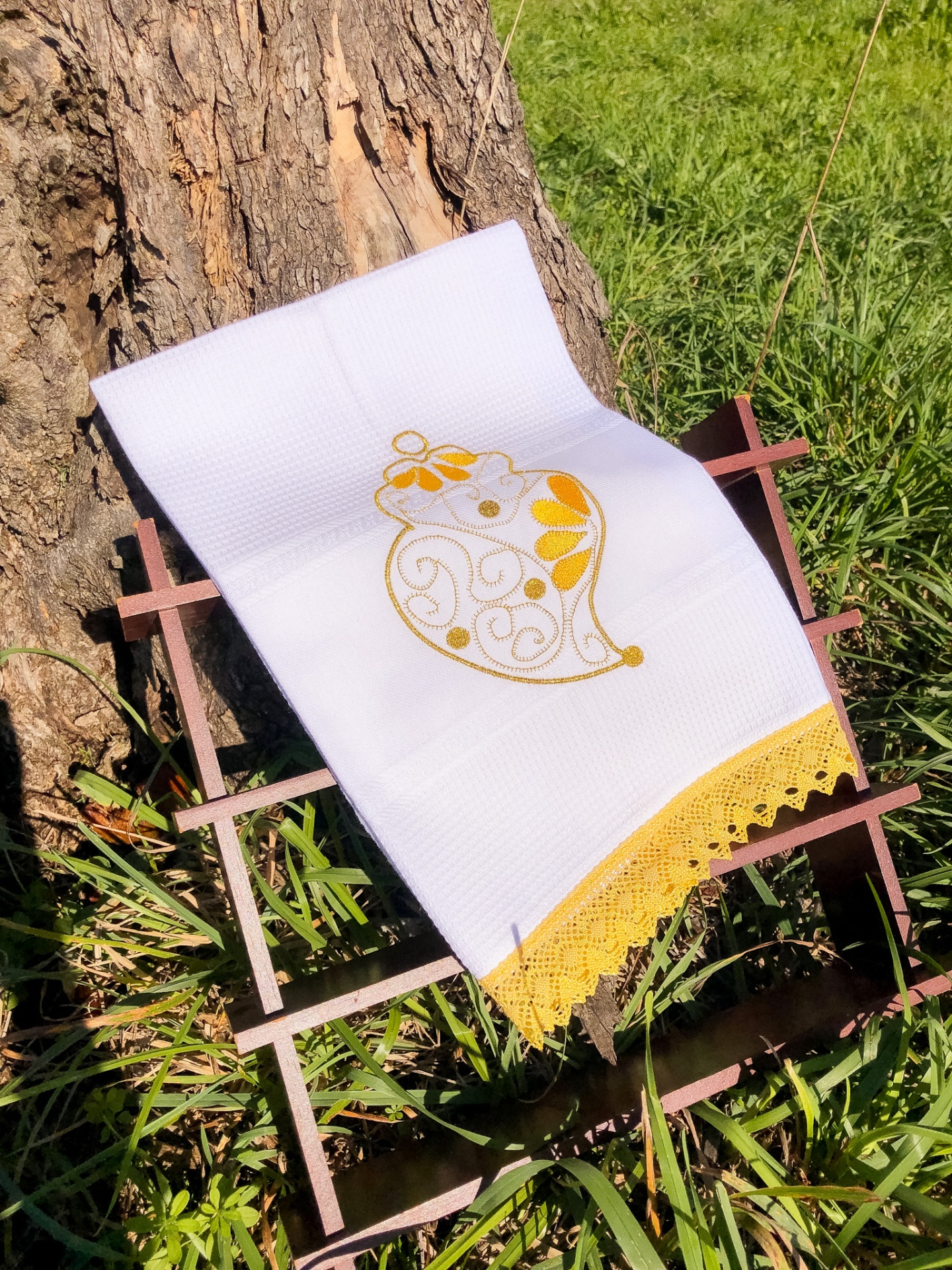 Toalha branca bordada com sino amarelo e renda amarela sobre suporte de madeira ao ar livre