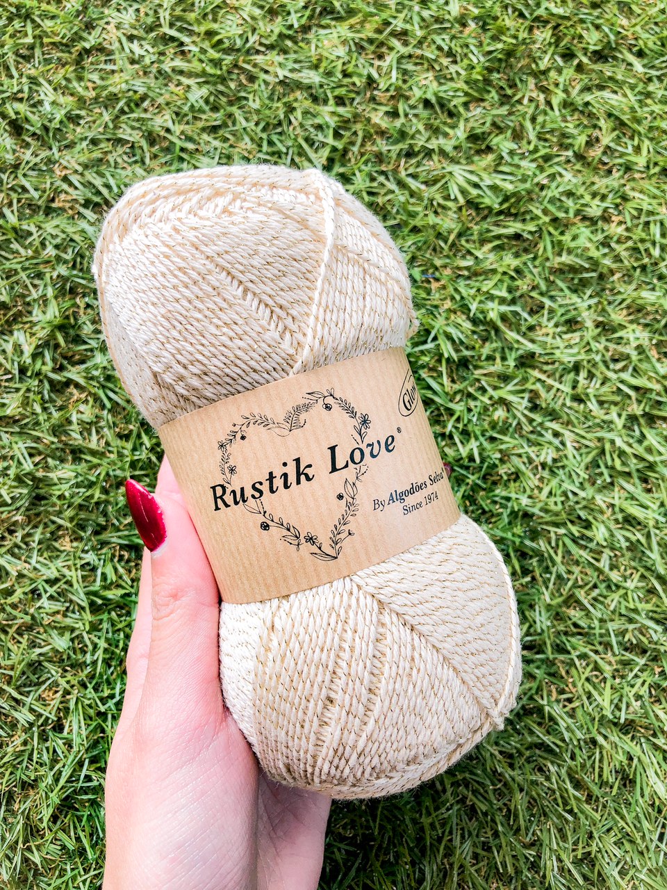Novelo de lã bege com rótulo Rustik Love, segurado por mão sobre relva verde