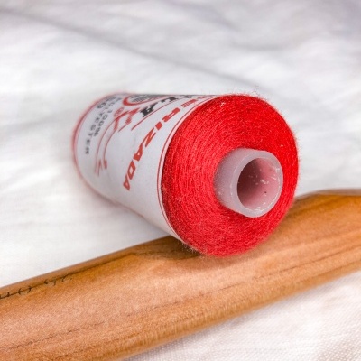 Rolo de linha vermelha em cima de tecido branco junto a objeto de madeira clara