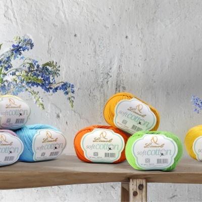 Novelos de lã Soft Cotton de várias cores sobre tábuas de madeira com flores azuis ao fundo