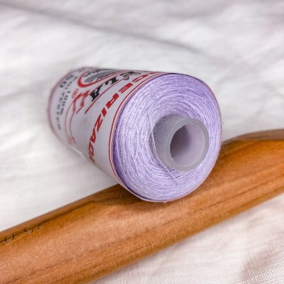 Carrete de linha lilás sobre tecido branco e cabo de madeira.