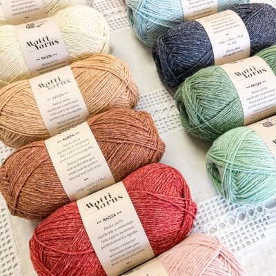 Várias bolas coloridas de lã etiquetadas Matti Yarns RAÍZZA em superfície branca rendada
