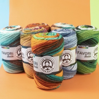 Seis novelos de fio multicolorido Favori Batik com etiquetas brancas sobre superfície alaranjada e fundo amarelo