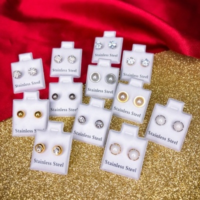 Conjunto de brincos em aço inoxidável com pedras, em suportes brancos, sobre fundo dourado e vermelho