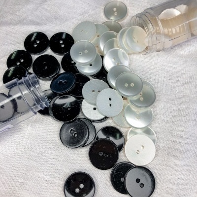 Botões pretos e brancos de dois furos espalhados sobre tecido branco, saindo de recipientes transparentes.
