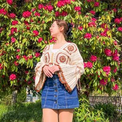 Mulher com blusa de malha bege e saia de ganga azul em frente a arbusto com flores cor de rosa