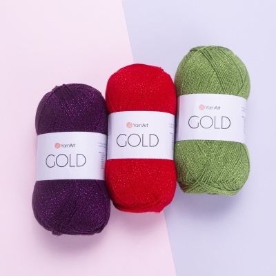 Três novelos de lã YarnArt GOLD em roxo, vermelho e verde sobre fundo rosa e lilás