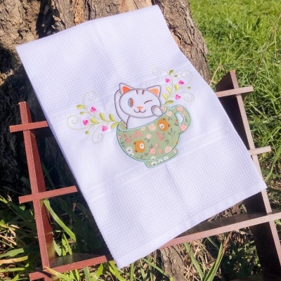 Toalha branca com bordado de gato numa chávena verde suportada por estrutura de madeira ao ar livre