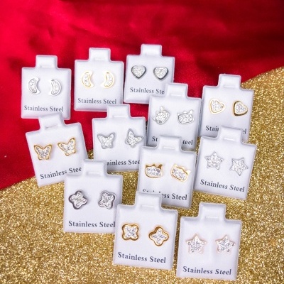 Conjunto de brincos de aço inoxidável em diversas formas e cores, em cartões brancos sobre tecido vermelho e dourado.