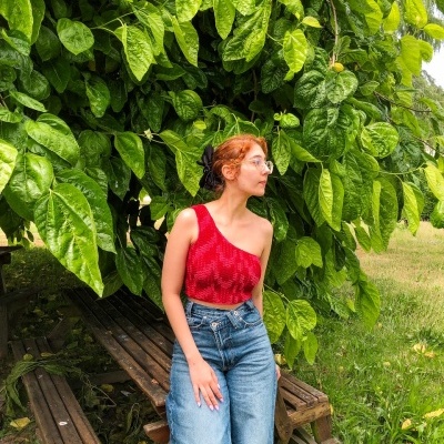 Mulher com top vermelho e calças de ganga sentada em banco de madeira junto a árvore com folhas grandes verdes
