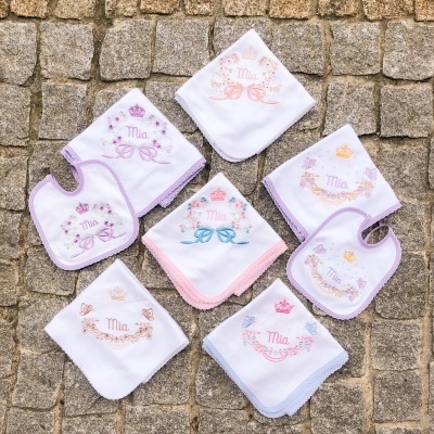 Babetes e fraldas brancas bordadas com nome Mia e motivos florais coloridos sobre calçada de pedra