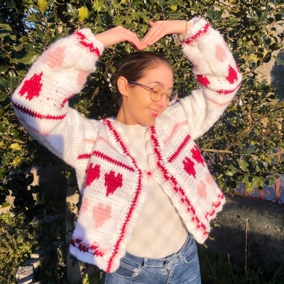 Mulher com casaco de malha branco com corações vermelhos e rosa, óculos, blusa branca e jeans, ao ar livre