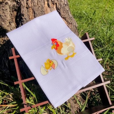Toalha branca com bordado de galinha e pintainho sobre estrutura de madeira junto a um tronco de árvore.
