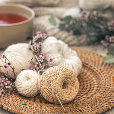 Novelos de lã bege e branco com agulhas de tricô num prato de rattan com flores lilases e chávena de chá ao fundo